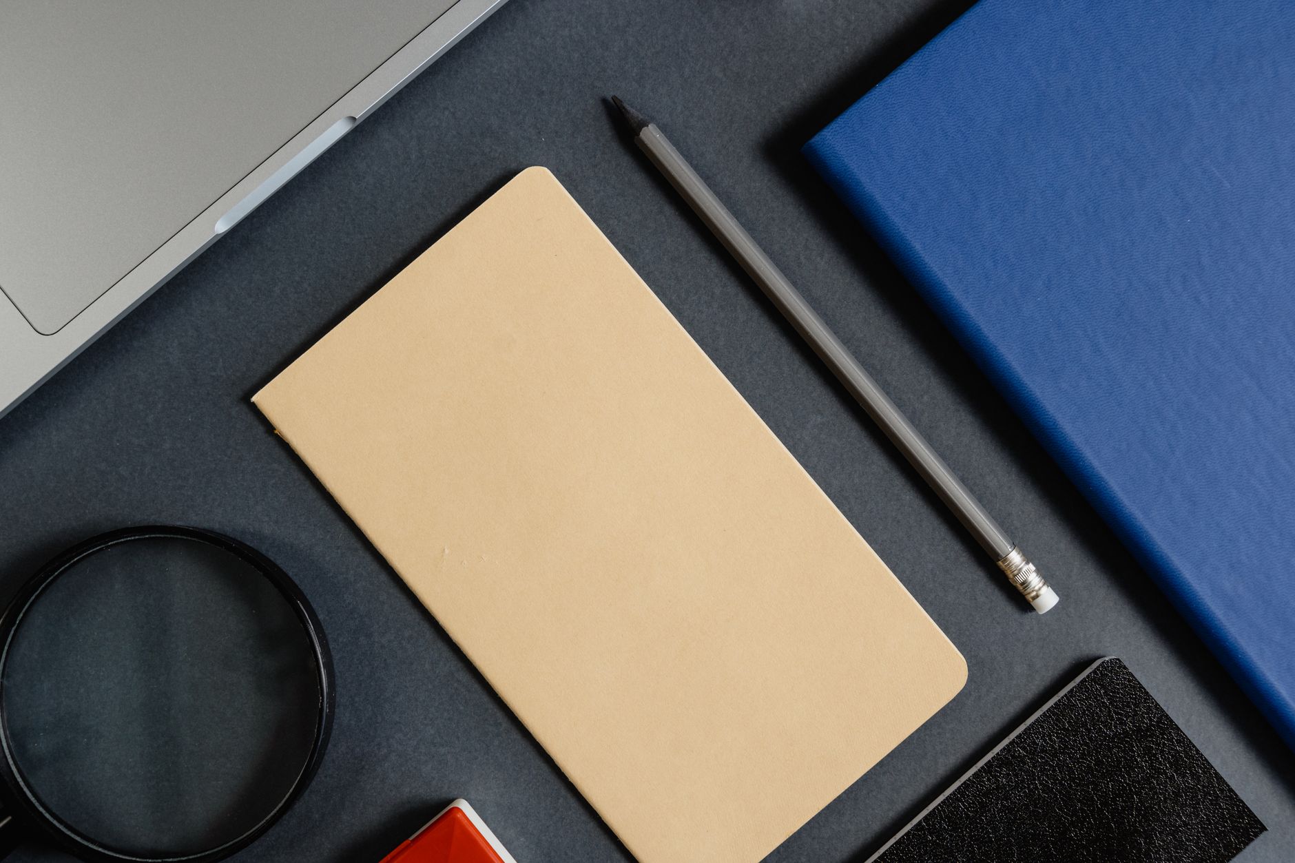 Vista aérea de uma mesa de trabalho escura com notebook, caderno azul, lápis e lupa, remetendo à rotina de quem presta serviços de escrita de casa.